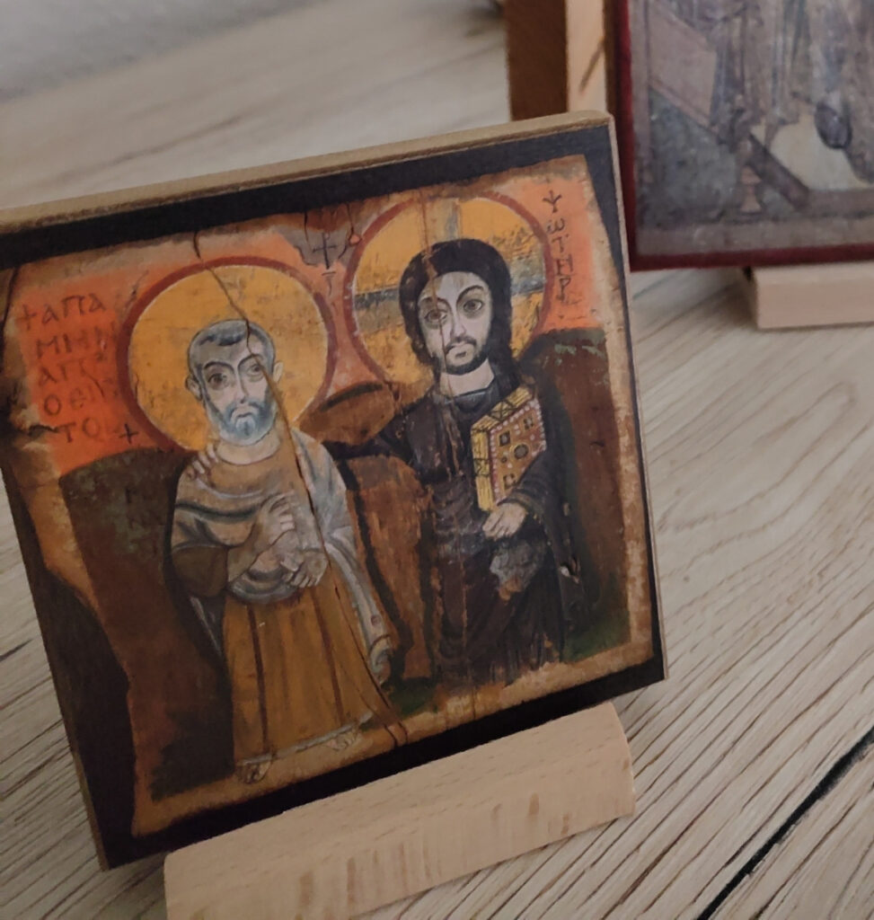 Ikone Christus und Abbas Menas: Jesus legt einer Person die Hand auf die Schulter, beide schauen nebeneinander aus dem Bild heraus. Im Hintergrund: Dreifaltigkeitsikone.